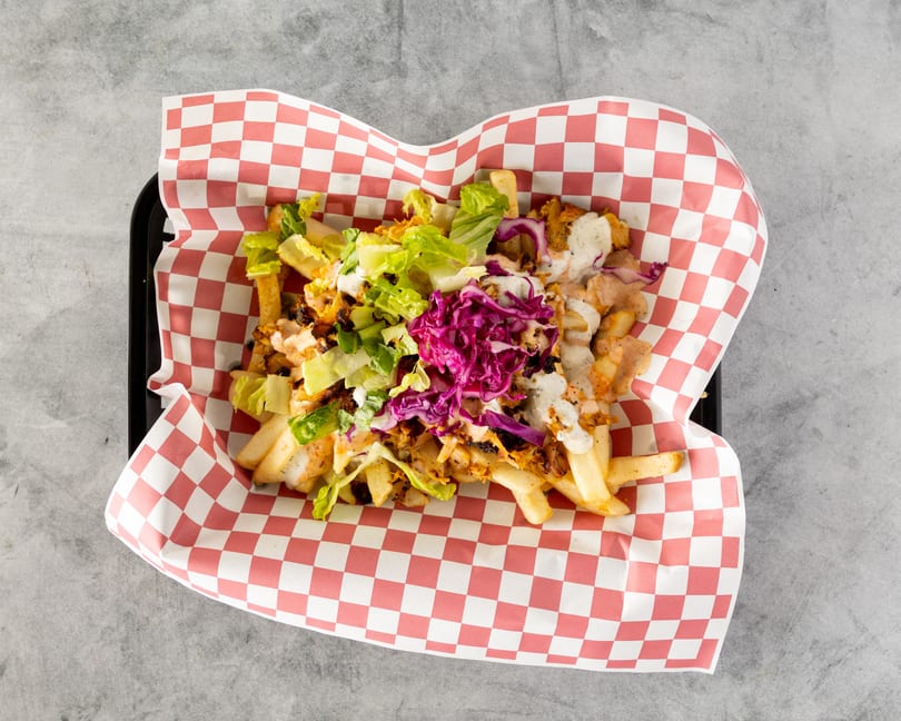 Loaded fries with chicken, lettuce, red cabbage, and two sauces in a red and white checkered paper.