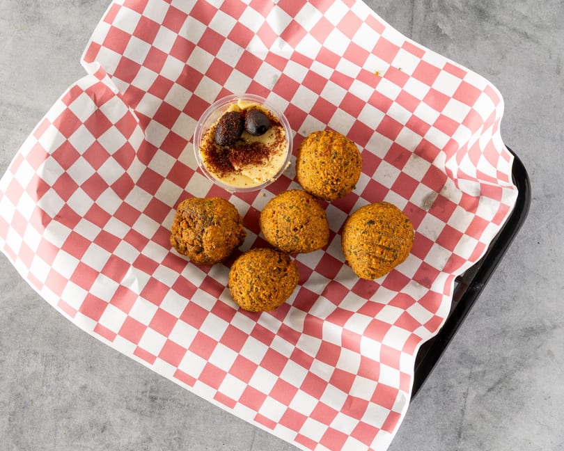Five falafel balls and a small container of hummus with olives on red and white checkered paper.