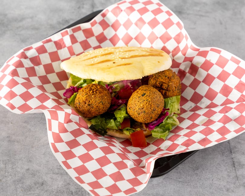 A falafel pita sandwich with lettuce, red cabbage, and tomatoes, served in a red and white checkered paper.