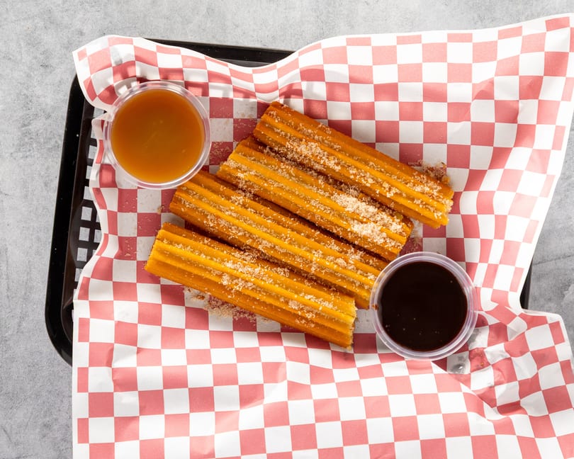 Churros sprinkled with sugar, served in a basket with red and white checkered paper, with caramel and chocolate dipping sauces.
