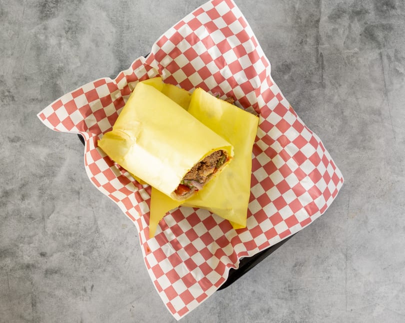 Two yellow-wrapped sandwiches with meat and vegetables, served in a red and white checkered paper-lined basket.